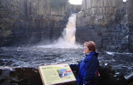 A woman stands by an information panel. There is a large waterfall behind her.