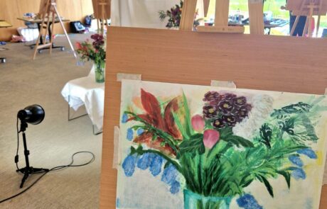 Photogrpah of artists easel with painting of vase of flowers on it and more easels in the background