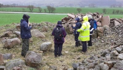 Photograph of group of people wearing waterproofs standing at a cairn of stones in a field