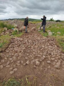 Photograph of two people standing at the far end of a strip of earth and stones uncovered in an archaeological dig