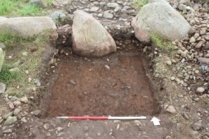 Photograph of archaeological dig with square hole, rule at front edge and large stones at the back