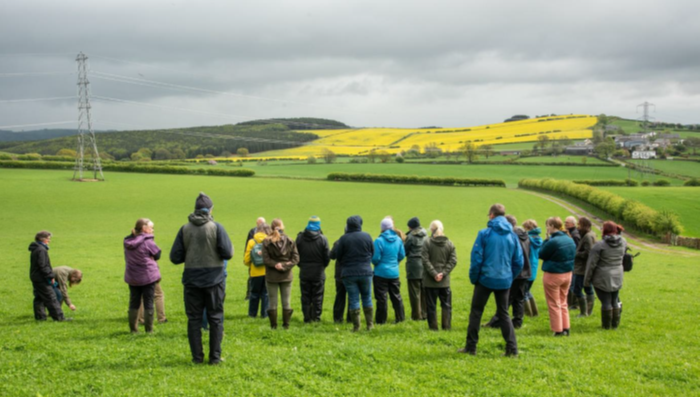 The image shows a group of people in the foreground standing in a field. In the background there are fields, hedges, hills and woodland. The sky is cloudy.