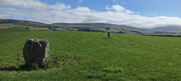 The image shows a field with a stone in the foreground and fells in the background. In the middle of the field there is a person carrying out a survey. 