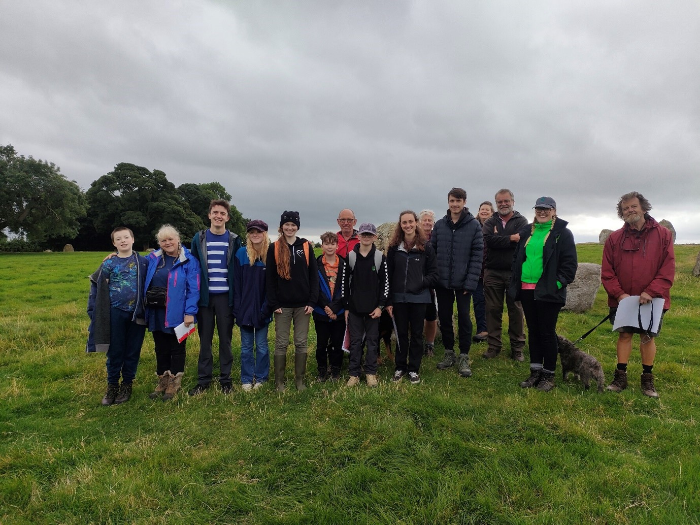 The image shows the group standing together in a field. There are some of the stones from the stone circle in the background. 