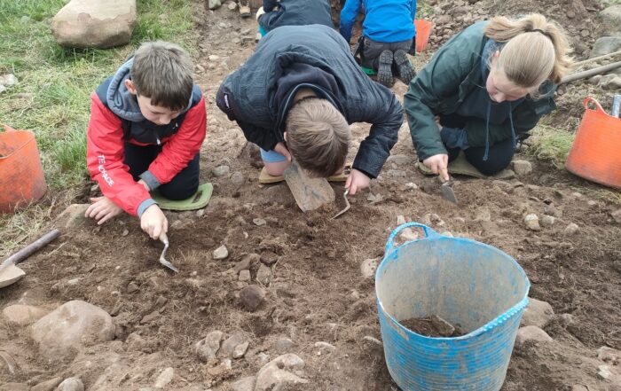 Students are kneeling on the ground. They are holding tools and scraping the ground. There is a bucket in the foreground with soil and rocks in. 2 students are also kneeling in the background with their backs to the camera.