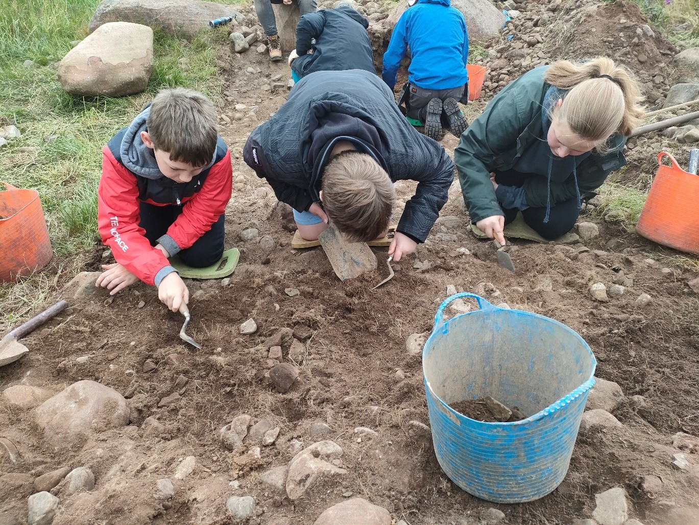 Students are kneeling on the ground. They are holding tools and scraping the ground. There is a bucket in the foreground with soil and rocks in. 2 students are also kneeling in the background with their backs to the camera. 