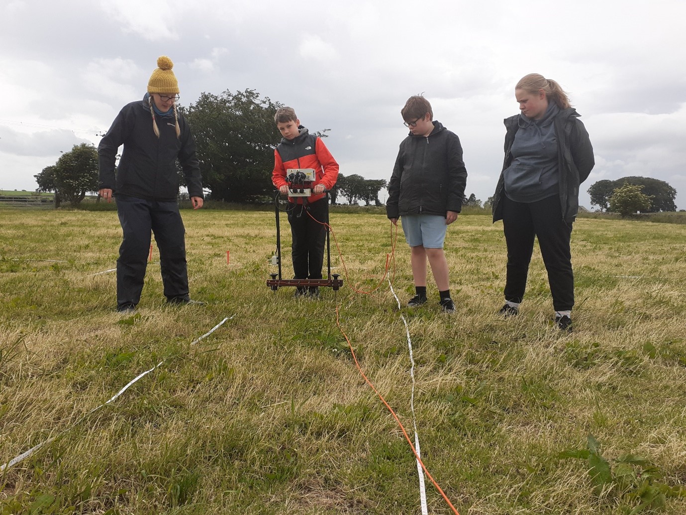Four people are standing in a field. One student holds a electrical resistivity tool as the other 3 watch. 