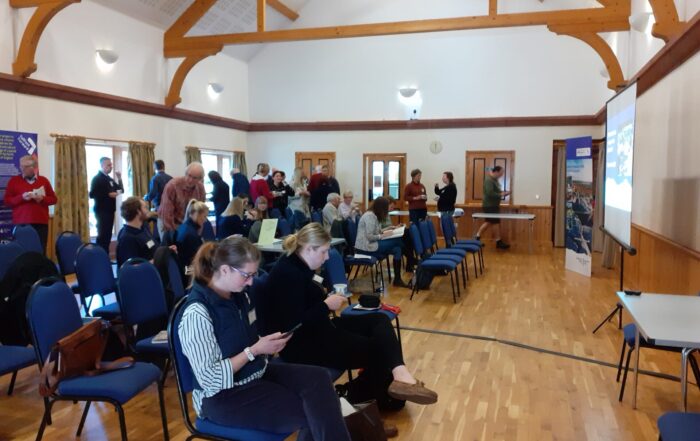 The image shows a village hall with people sitting o chairs in rows and others standing talking behind.