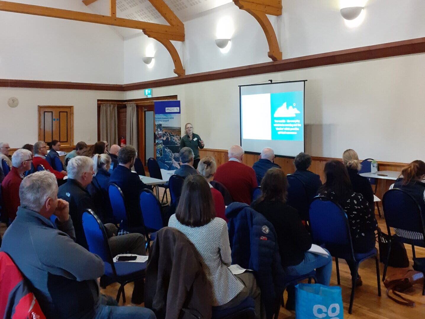 The image shows a village hall with the speaker standing in front of a projector screen. There are rows of people sitting in the audience.