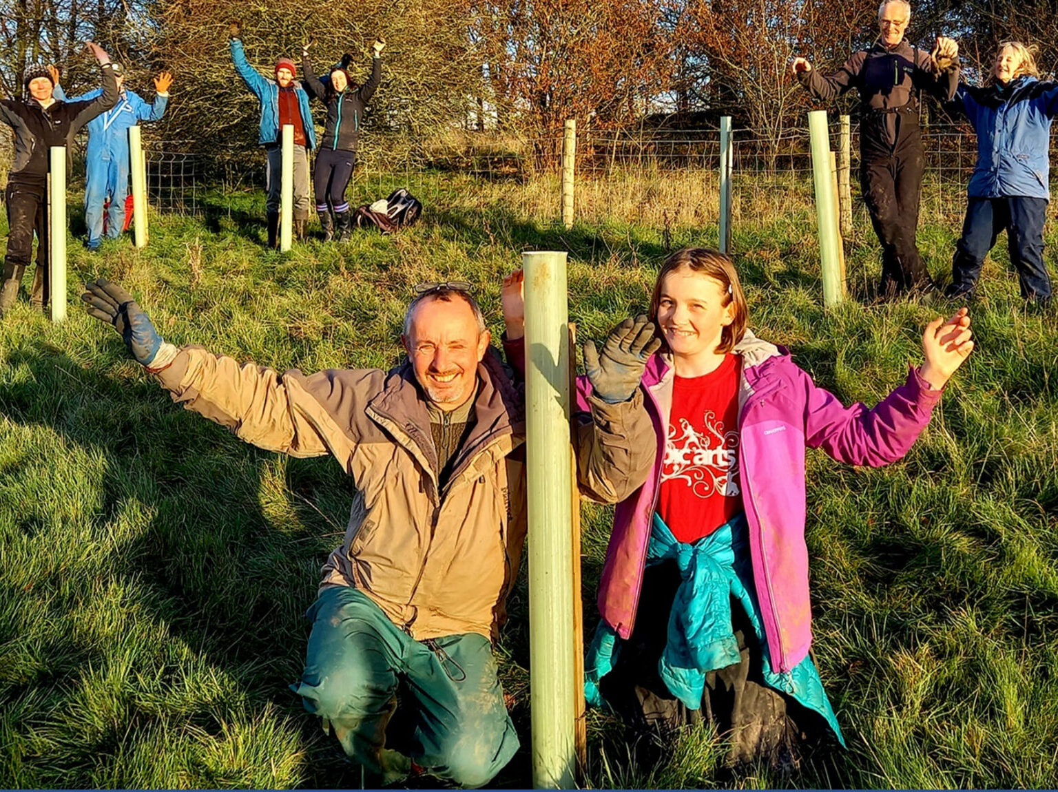 Photograph of adults and children in a field next to plastic tubes containing newly planted tree saplings