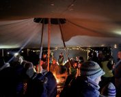 Adults dressed in winter clothes and sitting under large tent at stars and stories stargazing event. Fire in the centre.