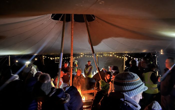 Adults dressed in winter clothes and sitting under large tent at stars and stories stargazing event. Fire in the centre.
