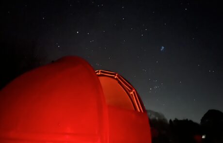 Alson Moor Observatory A red-illuminated observatory dome sits in the foreground under a dark, star-filled sky. The horizon is faintly visible, with silhouettes of trees and distant hills, while clusters of stars are bright and sharp against the night.