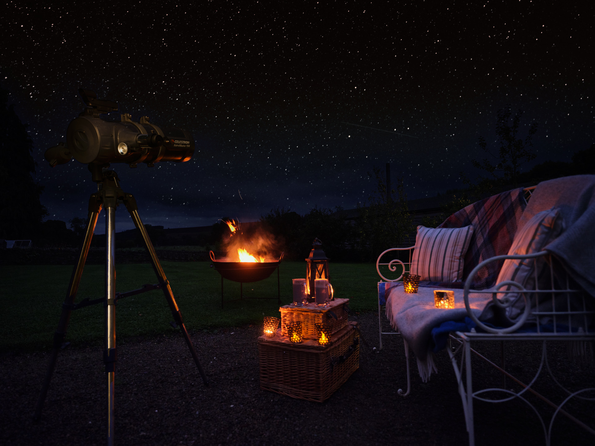 Outdoor stargazing scene at night: a telescope on a tripod faces a clear star-filled sky, while a fire pit glows in the background. In the foreground, woven baskets topped with lanterns and candles sit beside a cushioned bench draped with blankets and pillows, creating a warm, illuminated seating area under the stars.