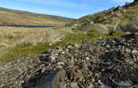 A heap of of flat,sharp pieces of stone in front of a grassy path and a wide river.